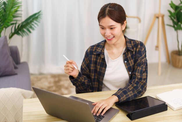 Levensstijl in woonkamerconcept Jonge Aziatische vrouw die op laptop typt en nota neemt van gegevens op tablet