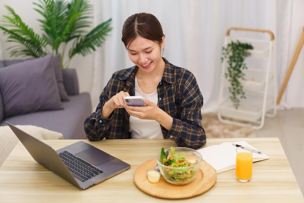 Levensstijl in woonkamerconcept Jonge Aziatische vrouw die smartphone gebruikt en groentesalade eet