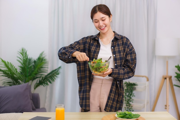 Levensstijl in woonkamerconcept Jonge Aziatische vrouw die zich bevindt om groentesalade in kom te mengen