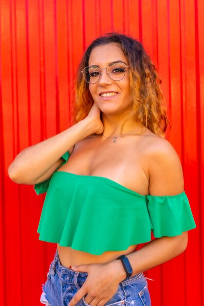 Levensstijl kaukasisch blond meisje in groene t-shirts en transparante bril op een rode muur lachend naar de zomerfotoshoot