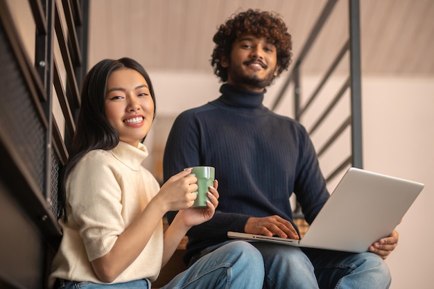 Foto levensstijl. mooie jonge aziatische vrouw met koffie en gekrulde bebaarde indiase man met laptop glimlachend zelfverzekerd kijkend naar camera zittend op trappen thuis