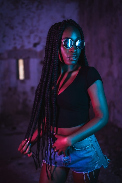Levensstijl portret van een jonge zwarte vrouw met lange vlechten zonnebril en een zwart t-shirt Roze en blauwe neonlichten stedelijke fotografie van een valdanser in ernstige pose