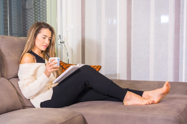 Levensstijl thuis, jonge blonde blanke vrouw die met een kopje koffie een boek leest op de bank in haar woonkamer