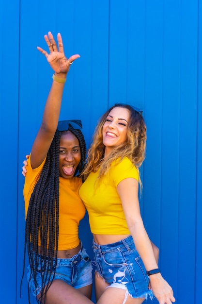 Levensstijl, tienermeisje vrienden genieten van in de fotoshoot op een blauwe muur gekleed in gele t-shirts. Zwart meisje met lange vlechten en blond Kaukasisch meisje.