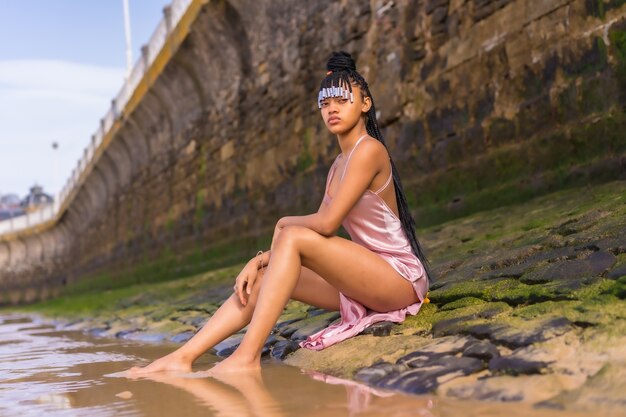 Levensstijl van een Dominicaanse etnische meisje met vlechten met een mooie roze jurk. Modieus poseren genietend van de zomer op het strand zittend op het zand