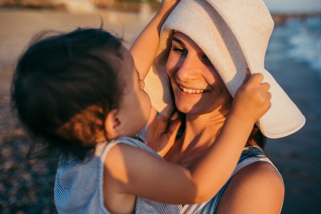 Levensstijl van jonge moeder spelen met dochter met witte hoed op de zee en strand achtergrond Familie vakantie Reizen Leuk vrouwelijk spel met vrolijke peuter blanke vrouw met baby op oceaan