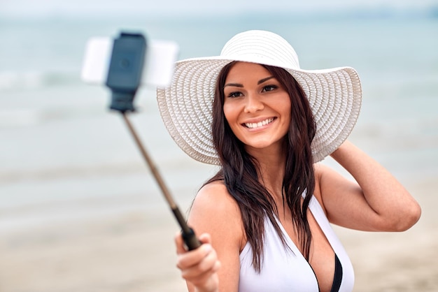 levensstijl, vrije tijd, zomer, technologie en mensenconcept - lachende jonge vrouw of tienermeisje in zonnehoed die foto's maakt met smartphone op selfiestick op het strand