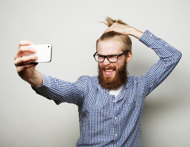 Levensstijlconcept een jonge man met een baard in hemd die een mobiele telefoon vasthoudt en een foto van zichzelf maakt terwijl hij tegen een grijze achtergrond staat