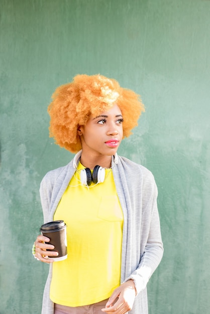 Levensstijlportret van een Afrikaanse vrouw in vrijetijdskleding die zich met koffie op de groene muurachtergrond bevindt