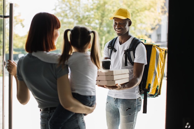 Levering afrikaanse man met thermisch geïsoleerde tas die pizzadoos en koffie geeft aan jong gezin