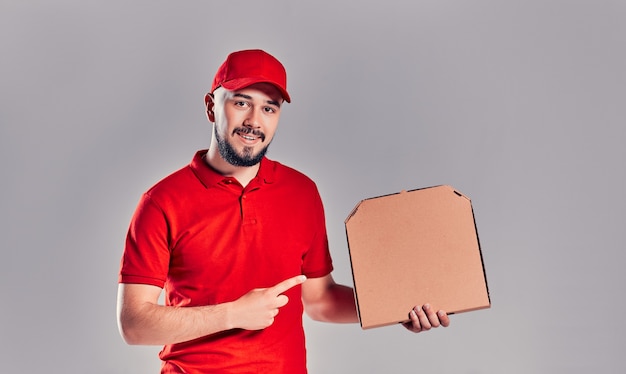 Levering man in rode dop, t-shirt geven eten bestellen pizzadozen geïsoleerd op een grijze achtergrond. mannelijke werknemer pizzaman koerier met pizza in lege lege kartonnen flatbox kopieerruimte. serviceconcept