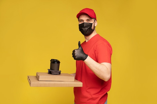 Levering man in rode pet, uniform t-shirt, beschermend masker en handschoenen geïsoleerd op gele muur