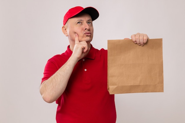 Levering man in rood uniform en pet houden papier pakket opzoeken met peinzende uitdrukking denken staande op witte achtergrond