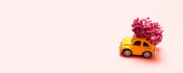 Levering. Stuk speelgoed gele auto met lila bloemtak op een roze eenvoudige achtergrond met plaats voor tekst.