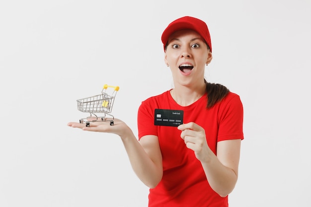 Levering vrouw in rode uniform geïsoleerd op een witte achtergrond. Vrouwelijke koerier of dealer in pet, t-shirt met supermarkt boodschappenwagentje om te winkelen, creditcard. Kopieer ruimte voor advertentie.