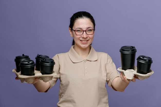 Levering vrouw werknemer in leeg poloshirt uniform dragen van een bril met koffiekopjes kijken naar camera gelukkig en positief glimlachend vrolijk staande over blauwe achtergrond