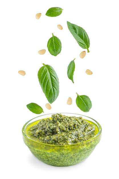 Levitating basil leaves and pine nuts above bowl of italian herbal pesto sauce isolated on white