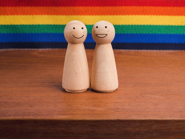 LGBT 커플 개념입니다. 치마 모양의 두 나무 인물, 행복한 미소 얼굴이 무지개 깃발 배경의 나무 탁자에 함께 서 있습니다. LGBT 프라이드 기호입니다.