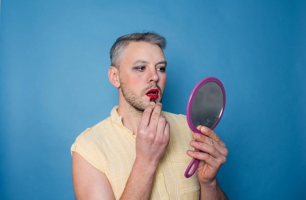 Лгбт-мужчина с макияжем на лице красит губы, глядя в зеркало на синем изолированном фоне