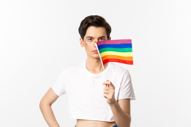 Lgbtq community. Attractive queer man with flitter on face, waving pride rainbow flag and looking at camera, standing in crop top against white.