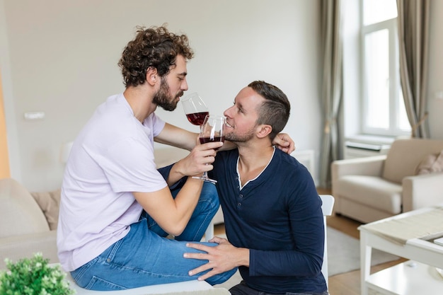 LGBTQ 커플 서로 껴안고 실내에서 와인을 마시며 거실에 함께 앉아 있는 동안 서로를 바라보는 두 로맨틱한 젊은 남성 연인 집에서 로맨틱한 젊은 게이 커플