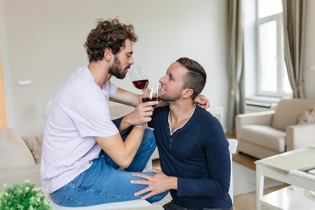 抱き合って室内でワインを飲む LGBTQ のカップル リビング ルームで一緒に座っている間、お互いを見ている 2 つのロマンチックな若い男性恋人 家でロマンチックな若い同性愛者のカップル