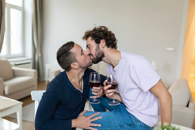 抱き合って室内でワインを飲む LGBTQ のカップル リビング ルームで一緒に座っている間、お互いを見ている 2 つのロマンチックな若い男性恋人 家でロマンチックな若い同性愛者のカップル