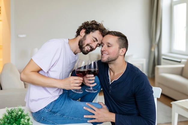 LGBTQ 커플 서로 껴안고 실내에서 와인을 마시며 거실에 함께 앉아 있는 동안 서로를 바라보는 두 로맨틱한 젊은 남성 연인 집에서 로맨틱한 젊은 게이 커플