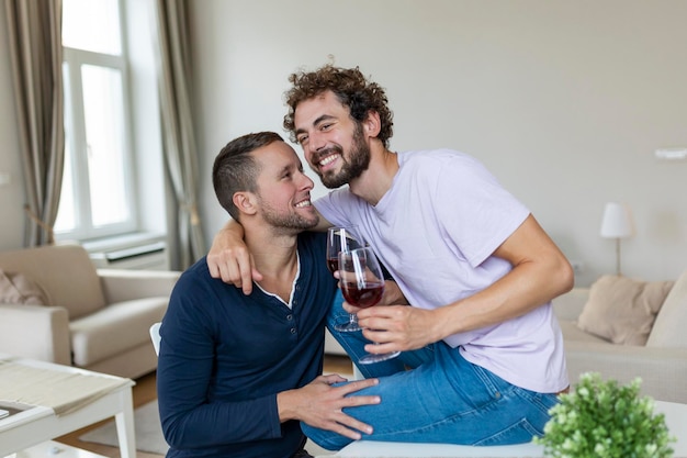 抱き合って室内でワインを飲む LGBTQ のカップル リビング ルームで一緒に座っている間、お互いを見ている 2 つのロマンチックな若い男性恋人 家でロマンチックな若い同性愛者のカップル