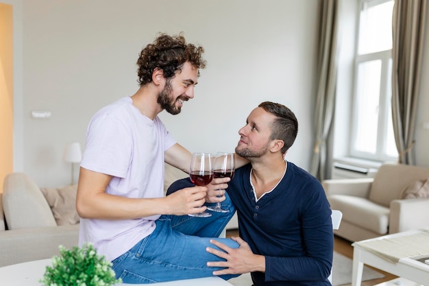 抱き合って室内でワインを飲む LGBTQ のカップル リビング ルームで一緒に座っている間、お互いを見ている 2 つのロマンチックな若い男性恋人 家でロマンチックな若い同性愛者のカップル