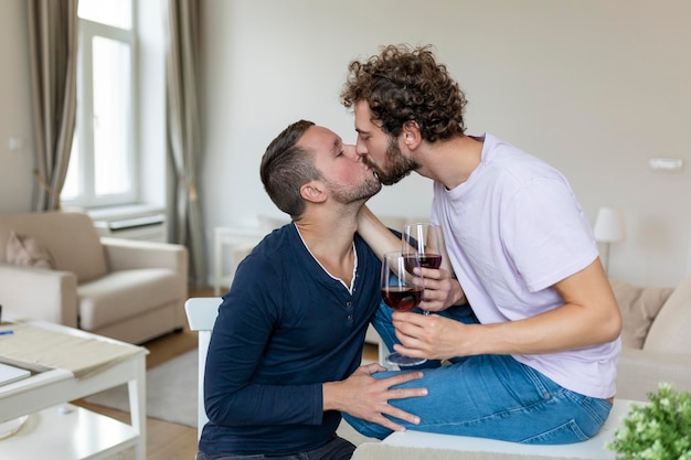 Coppia lgbtq che si abbracciano e bevono vino al chiuso due romantici giovani amanti maschi che si guardano mentre si siedono insieme nel loro soggiorno giovane coppia gay che è romantica a casa