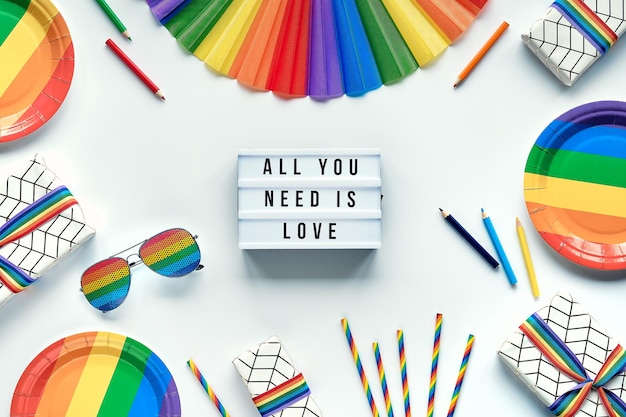 LGBTQ pride month decor. Text All You Need Is Love on lightbox. Rainbow pattern objects on off white background. Party paper plates, drink straws, sunglasses and fan. Flat lay, top view.