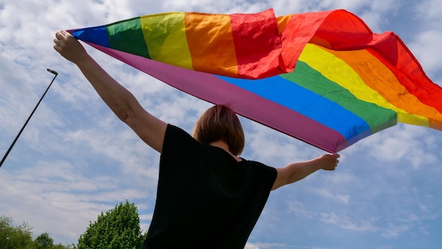 Lgbtqia의 달 프라이드 무지개 평화 깃발은 화창한 날 구름과 함께 푸른 하늘을 배경으로 한 여자를 안고 있다