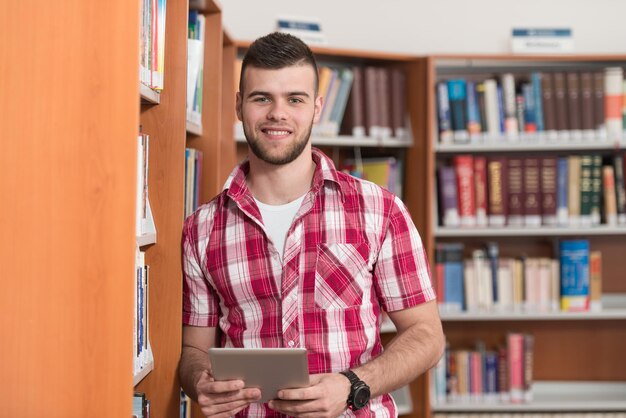 図書館でノートパソコンと本を持ったハンサムな男子生徒が高校の大学図書館で働いている浅い被写界深度