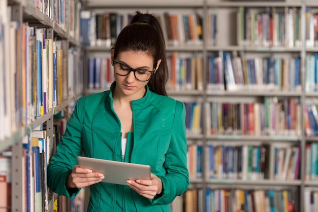 В библиотеке красивая студентка с планшетом и книгами, работающая в библиотеке старшей школы, малая глубина резкости