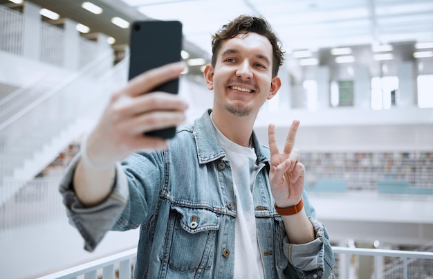 Foto selfie della biblioteca e uomo dell'università con segno di pace per aggiornare lo stile di vita social media o post sul blog con felice educazione studio di ricerca e dottorando o persona con segno emoji per l'immagine del profilo