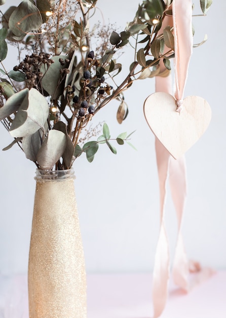 Licht houten hart hanger aan een roze zijden lint op een achtergrond van een gouden glanzende glittervaas met gedroogde eucalyptus takken.