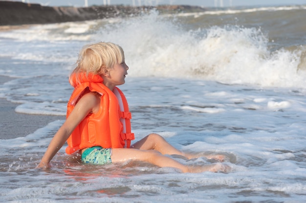 Lichtharige jongen in een oranje opblaasbaar vest zittend aan de kust en wachten op grote golf.