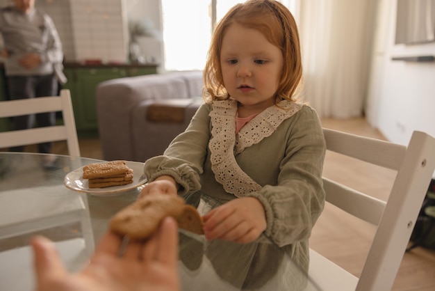 Lichthuidig roodharig meisje in jurk kijkt naar koekjes terwijl ze aan tafel zit. Voedingsconcept voor kinderen