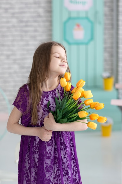 Lief klein donker haarmeisje in purpere kleding met gele tulpen in studio