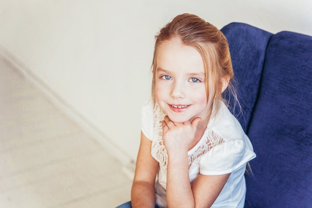 Lief klein meisje in spijkerbroek en wit T-shirt zittend op blauwe stoel thuis ontspannen