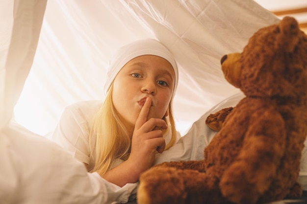 Lief klein meisje met een teddybeer houdt de vinger op de lippen en toont stilte om een geheim te bewaren terwijl ze op haar bed ligt in een tent met slaapmutsje