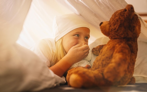 Lief klein meisje speelt met een teddybeer en houdt hand voor haar mond om een geheim te bewaren terwijl ze op haar bed ligt in een tent met slaapmutsje