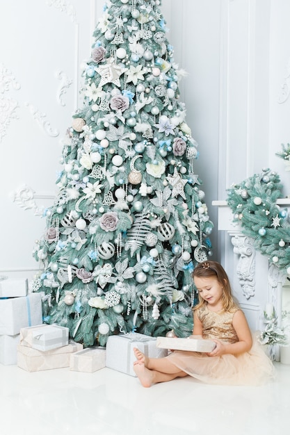 Lief meisje zit in de buurt van een kerstboom in de kamer en overweegt geschenken.