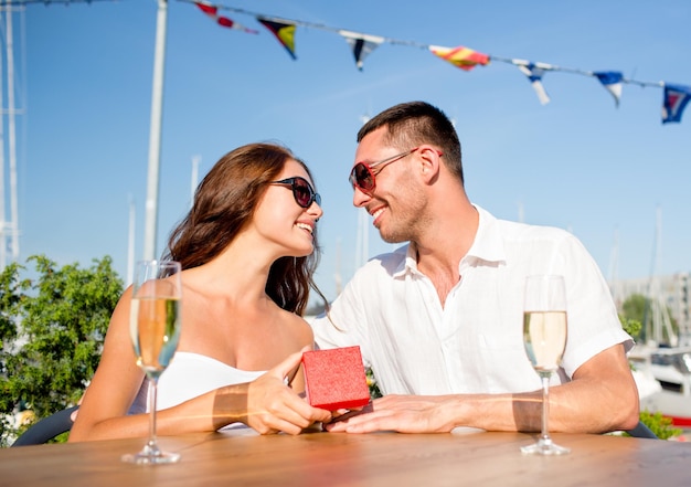 liefde, dating, mensen en vakantie concept - glimlachend paar met zonnebril met champagne en rode geschenkdoos die naar elkaar kijkt in café