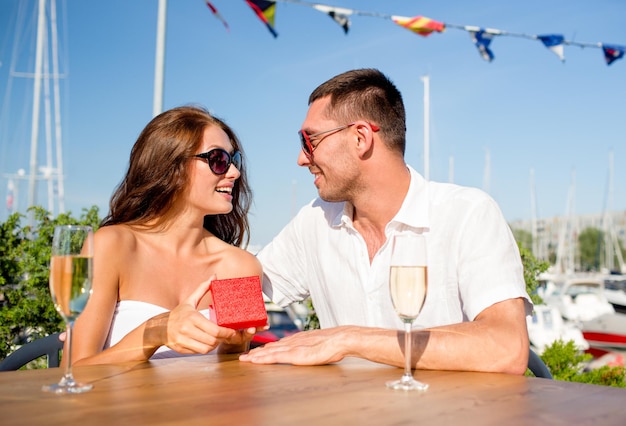 liefde, dating, mensen en vakantie concept - glimlachend paar met zonnebril met champagne en rode geschenkdoos die naar elkaar kijkt in café