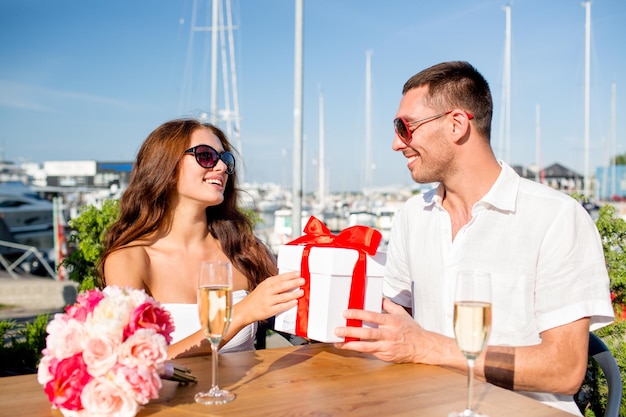 liefde, dating, mensen en vakantie concept - glimlachend paar met zonnebril zittend met geschenkdoos, bloemen en champagne in café