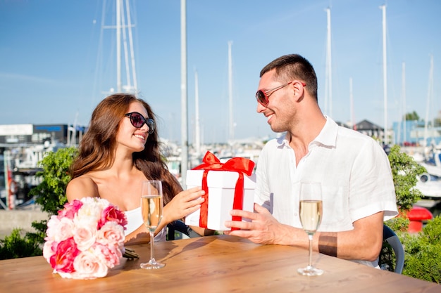 liefde, dating, mensen en vakantie concept - glimlachend paar met zonnebril zittend met geschenkdoos, bloemen en champagne in café