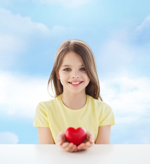 liefde, liefdadigheid, jeugd en mensen concept - mooi klein meisje zittend aan tafel en met rood hart over bewolkte hemelachtergrond