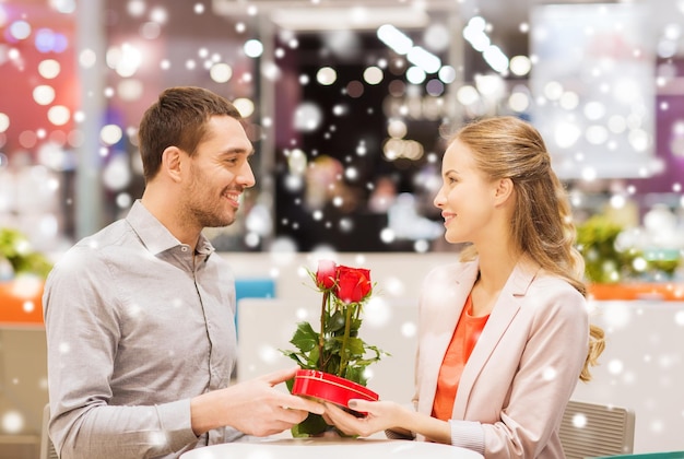 liefde, romantiek, Valentijnsdag, paar en mensen concept - gelukkige jonge man met rode bloemen cadeau geven aan lachende vrouw in café in winkelcentrum met sneeuweffect